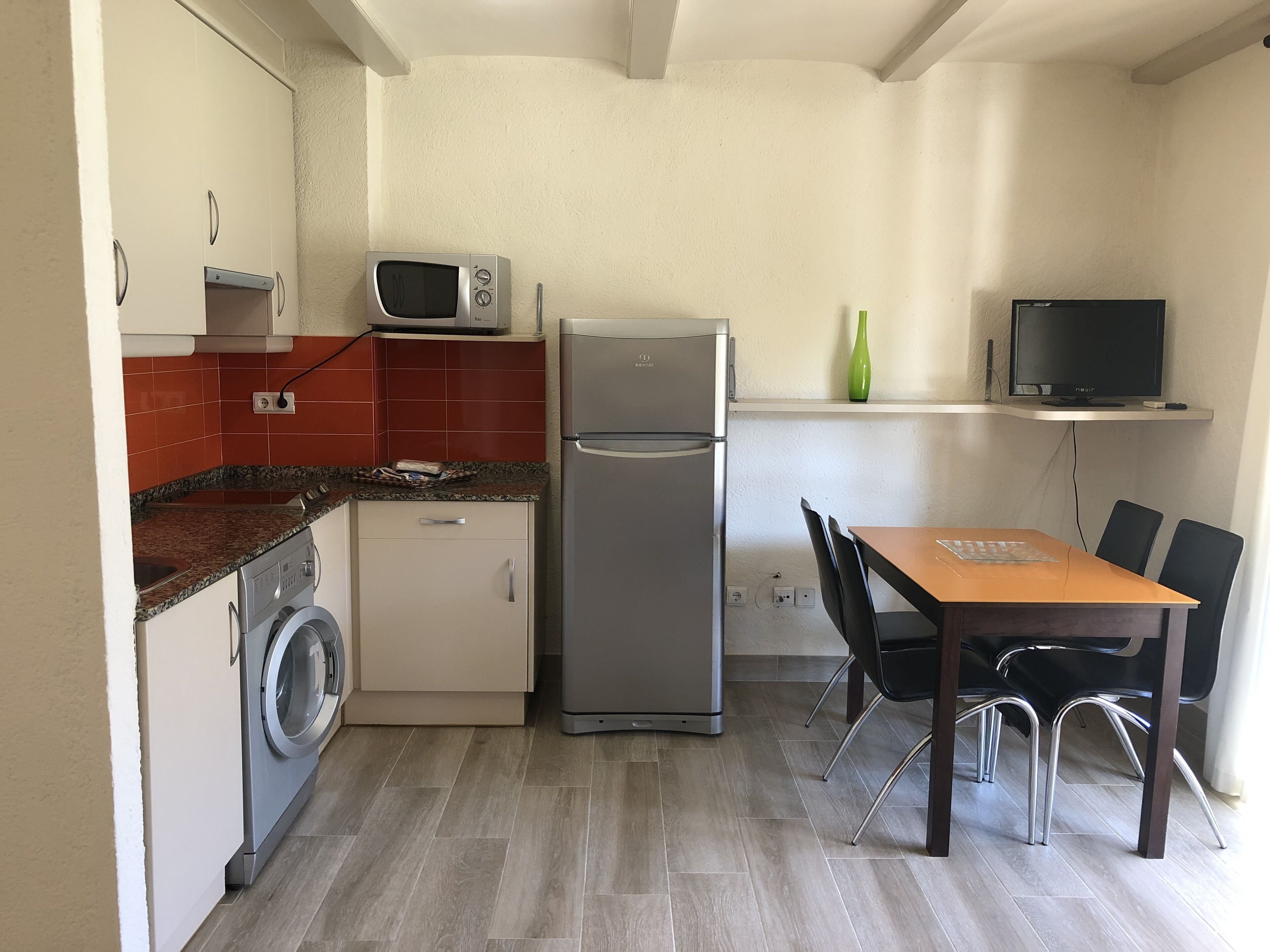 A compact kitchen with a washing machine, a refrigerator, a microwave, and a dining area with a table and chairs.