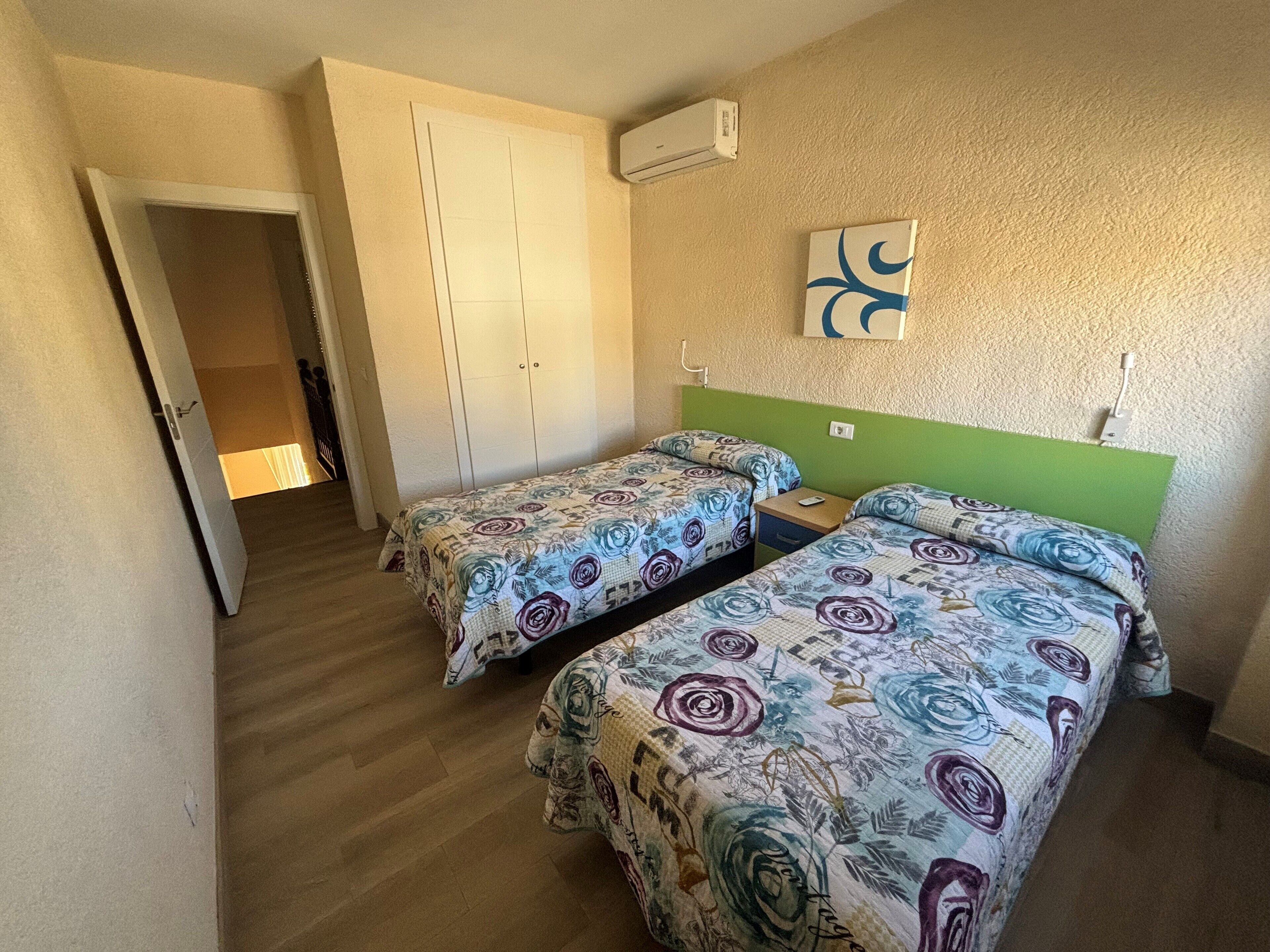 A room with two single beds, a wardrobe, an air conditioning unit, and a small table.