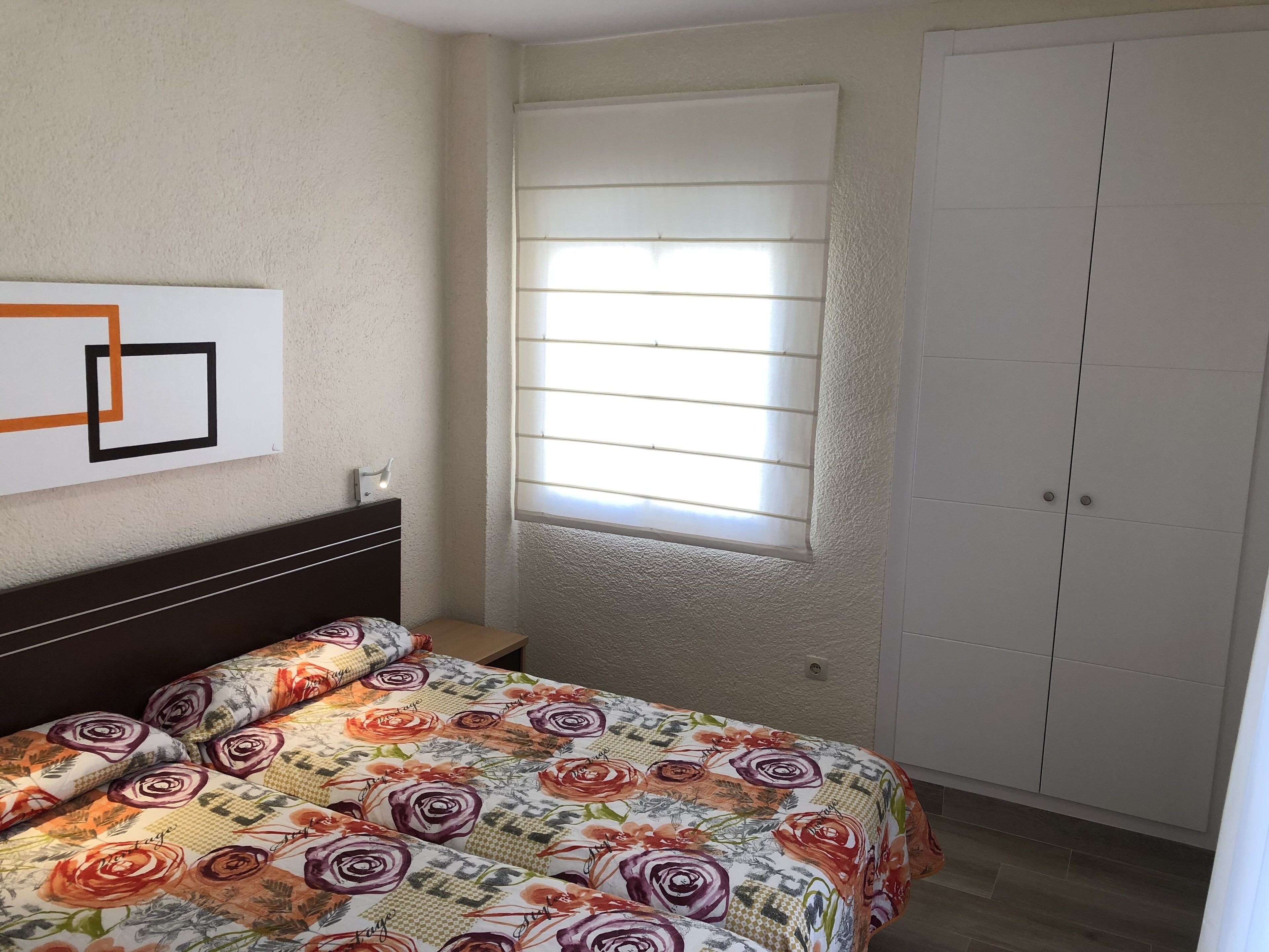 A bedroom with a bed, a window with blinds, a wardrobe, and a wall-mounted light fixture.