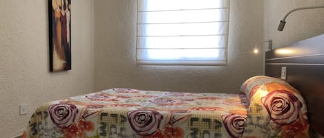 A bedroom with a bed, a window with blinds, a painting on the wall, and a bedside lamp.
