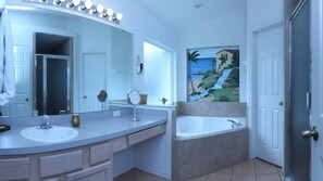 A bathroom with a large mirror, two sinks, a bathtub, and a painting of a coastal landscape.