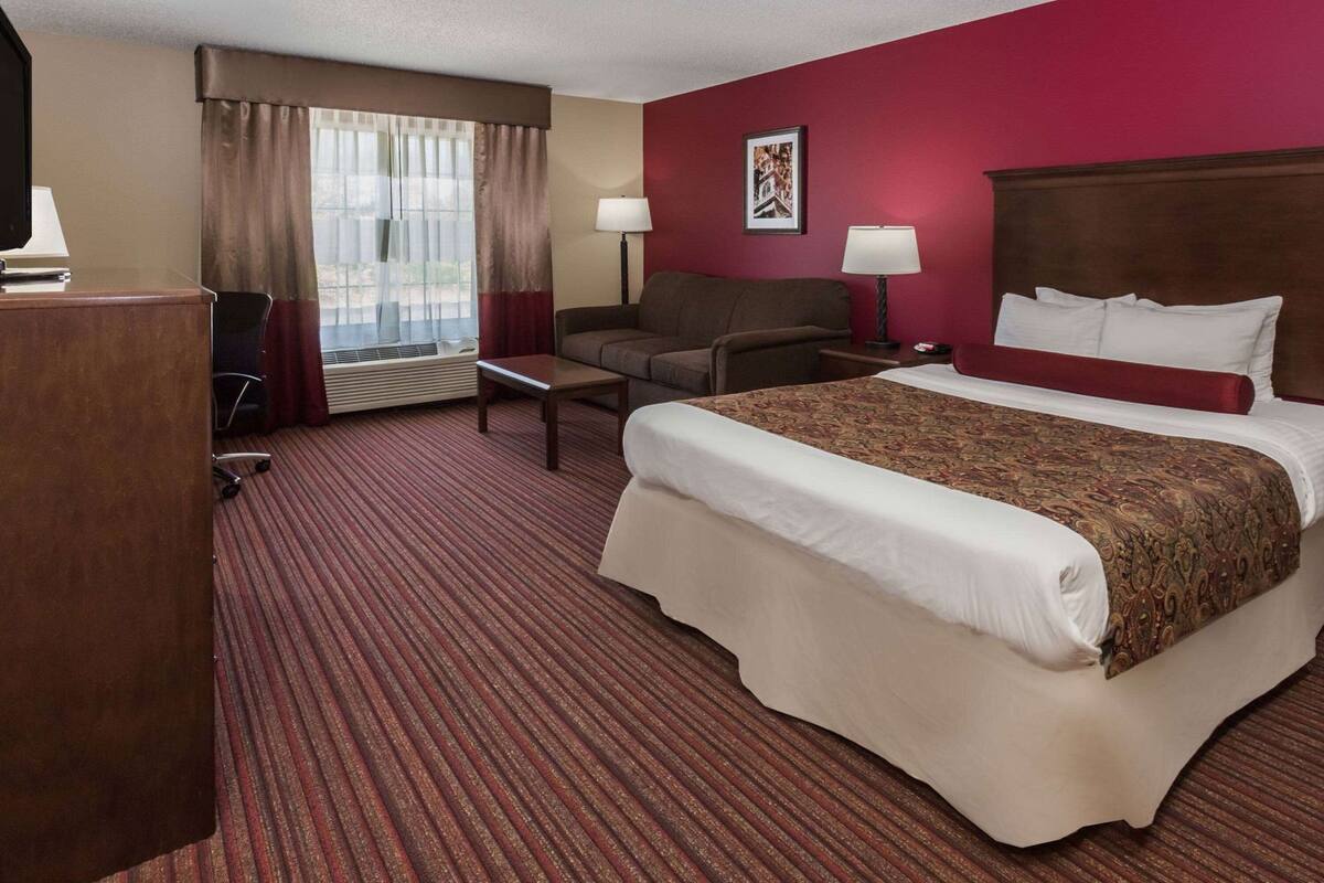 Ein Hotelzimmer mit einem großen Bett, einem Schreibtisch, einem Stuhl, einer Couch, einem Fenster mit Vorhängen und einem Wandbild.