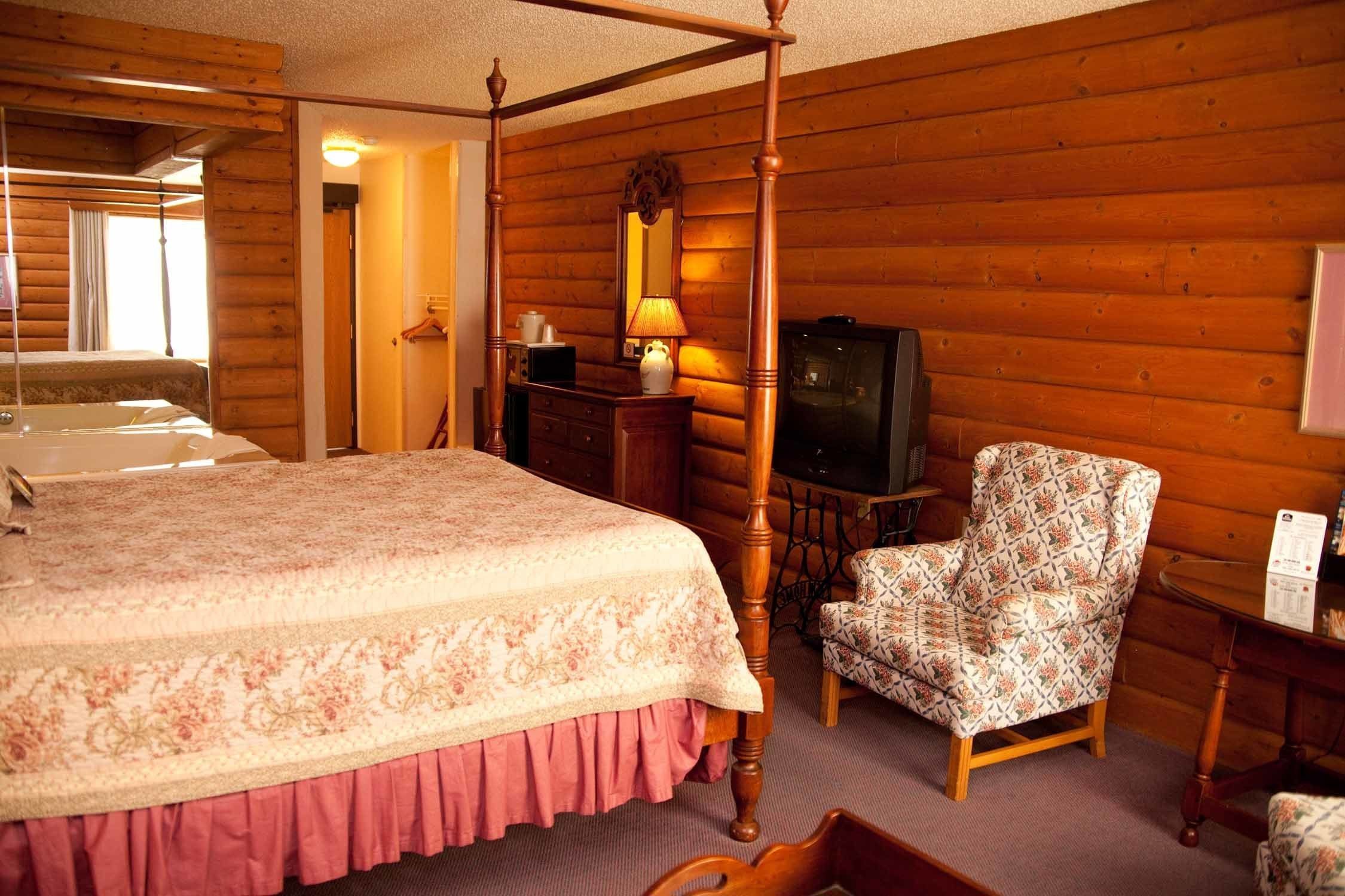 Un dormitorio de estilo cabaña de troncos con una cama con dosel, un aparador de madera, un sillón estampado y un televisor.