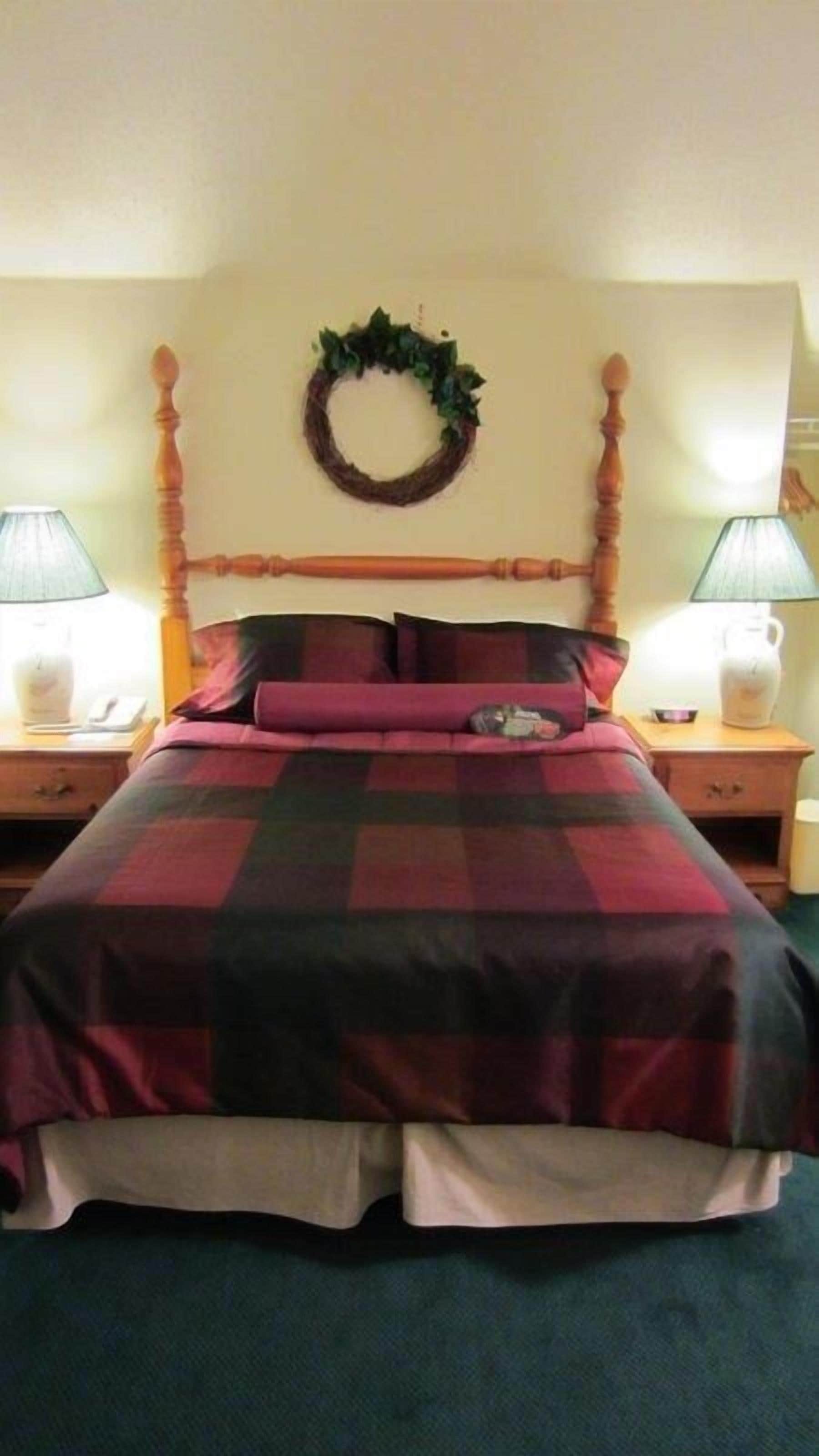 Een bed met een geruite sprei, een houten hoofdbord en een krans aan de muur. Er staan twee nachtkastjes met lampen.