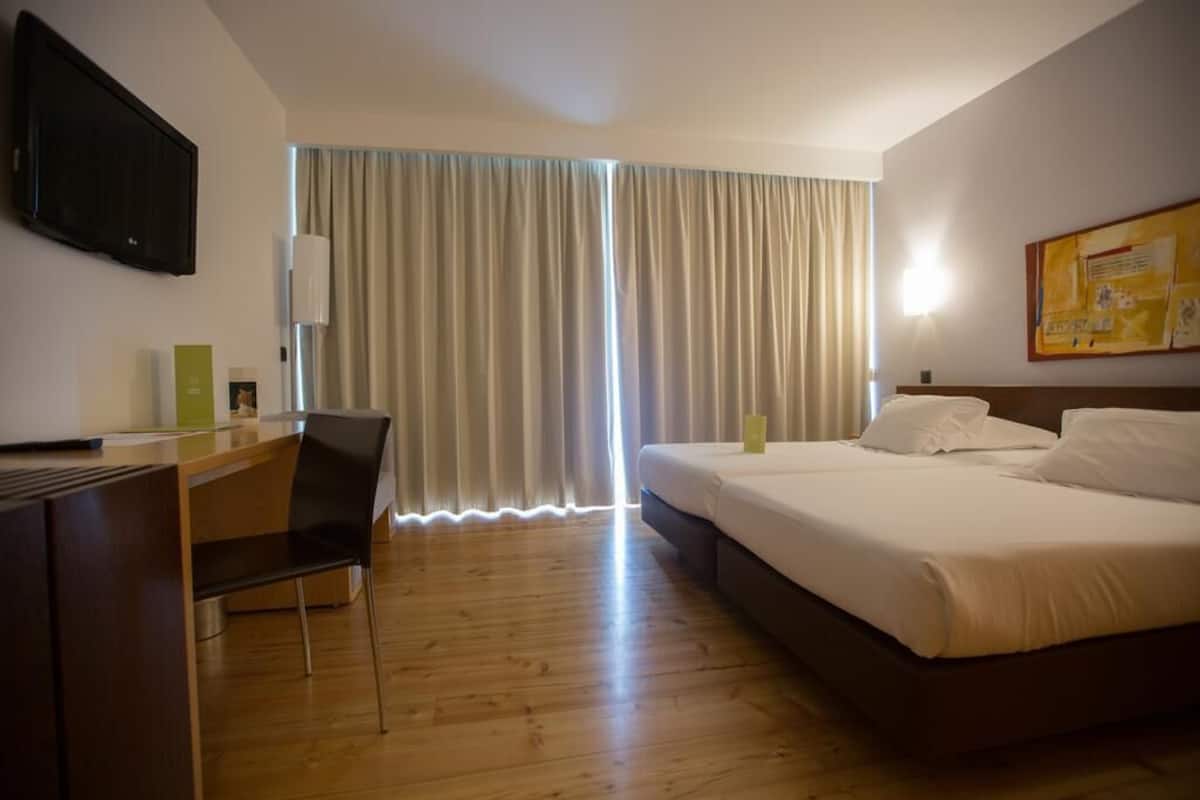 Habitación de hotel con piso de madera, un escritorio con silla, cama con ropa blanca y un ventanal con cortinas.