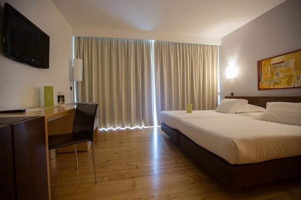 Habitación de hotel con piso de madera, un escritorio con silla, cama con ropa blanca y un ventanal con cortinas.