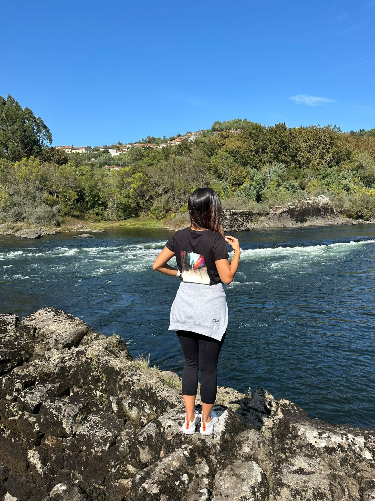 Una persona parada sobre rocas junto a un río, con un paisaje boscoso en el fondo.
