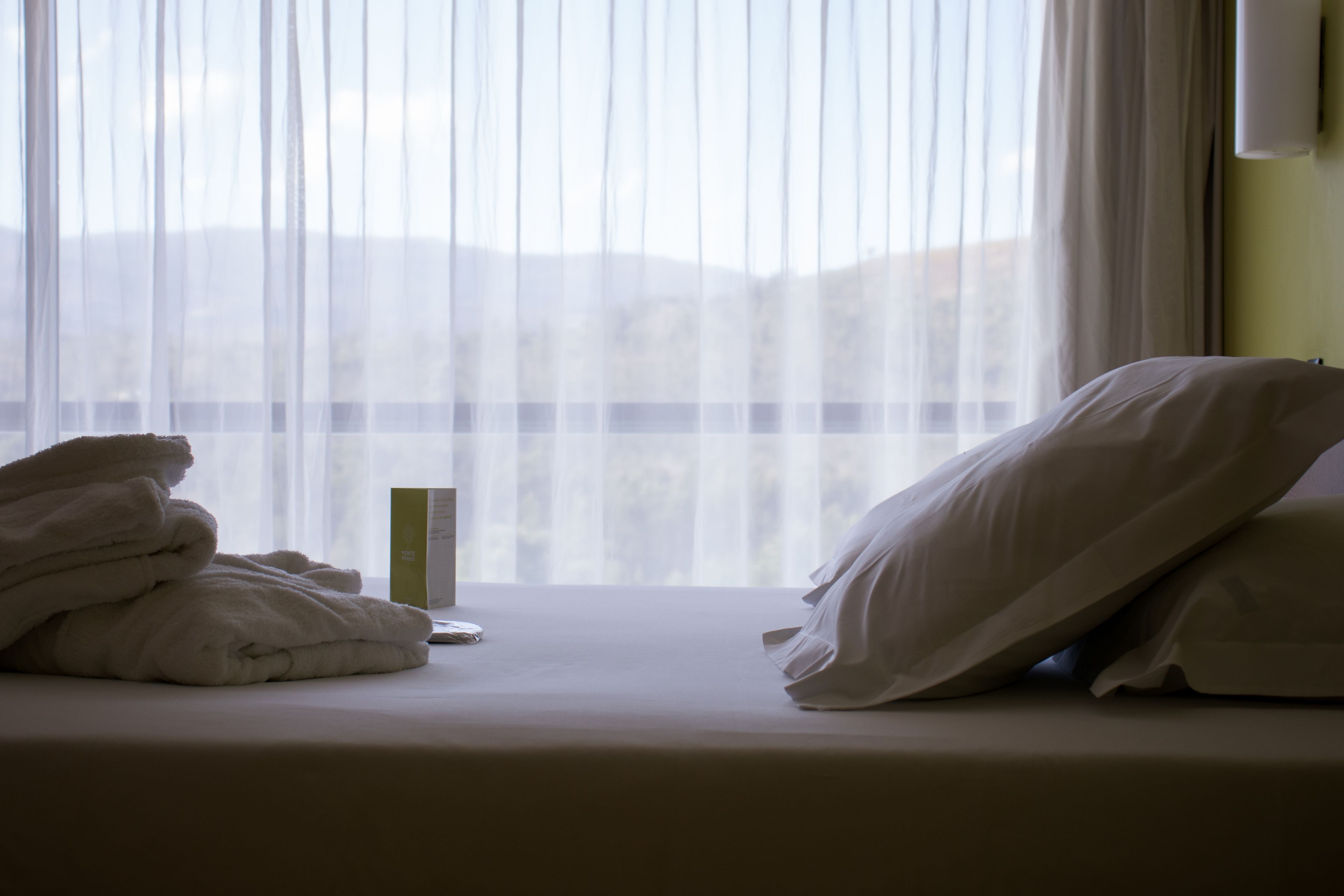 Una habitación de hotel con cama, ropa de cama blanca, mesita de noche y ventana con cortinas de gasa.
