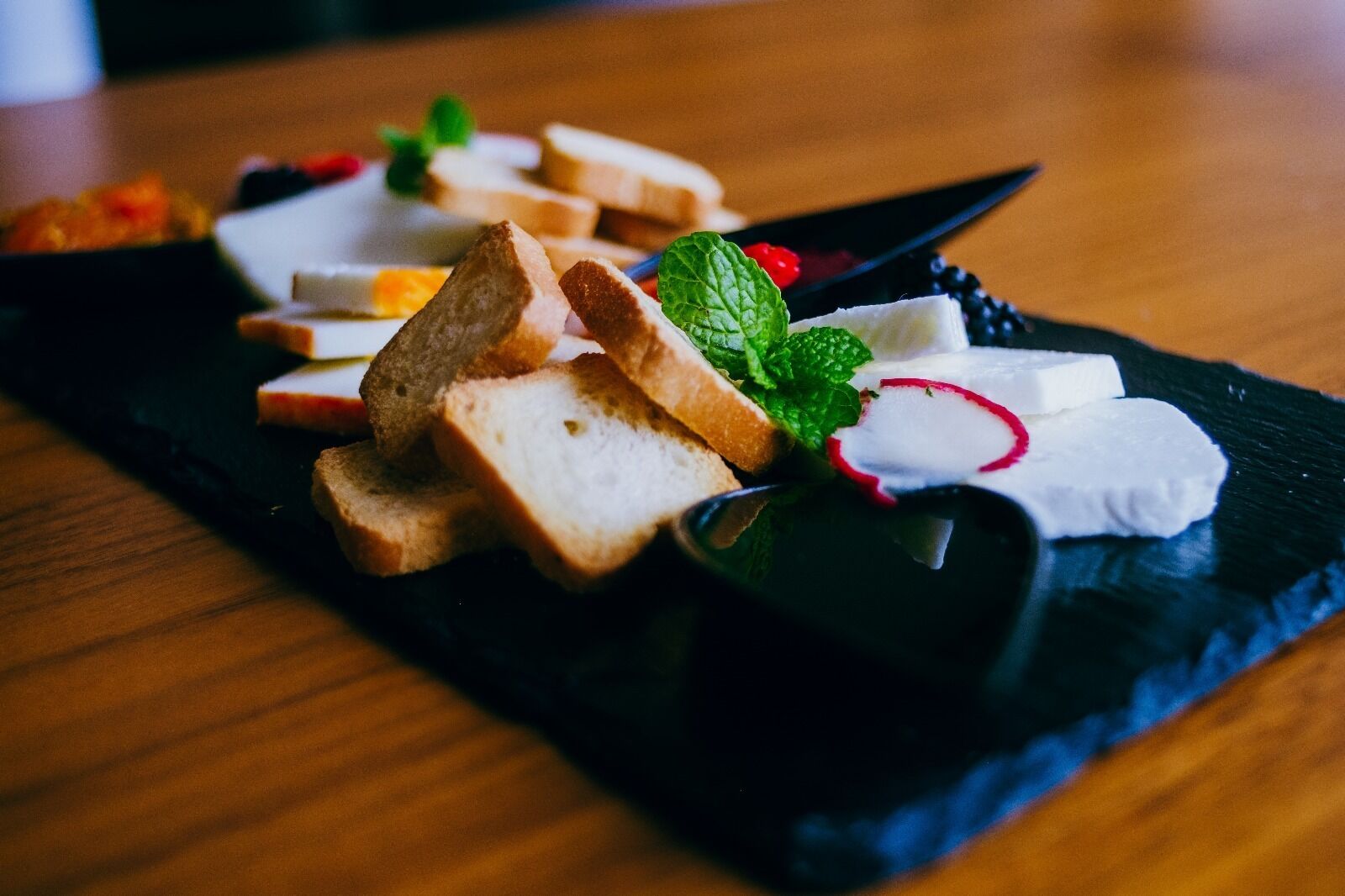 A black slate serving board with slices of bread, cheese, radishes, and mint leaves.