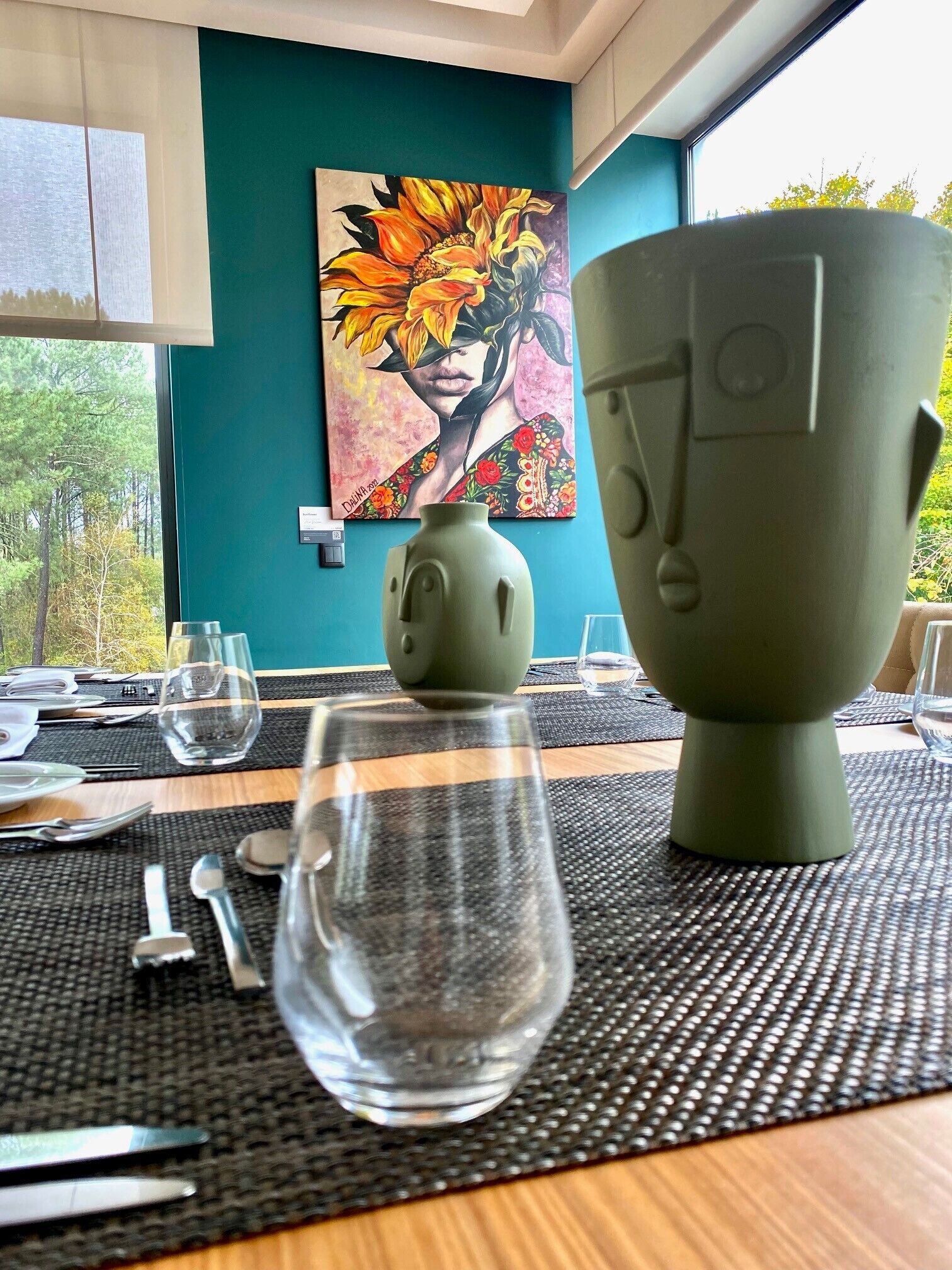 A dining table with a vase, a large cup, and a glass, set against a teal wall with a painting.