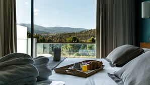 A hotel room with a large window offering a mountain view, a bed with pillows, a tray with breakfast items, and a bedside table.