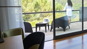 A spacious room with a large window offering a view of a lush landscape. There is a balcony with a railing, and a person is visible standing on it.