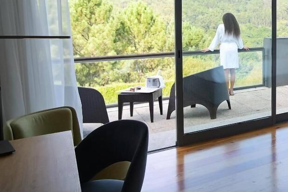 Amplia habitación con un ventanal que ofrece vistas de un paisaje exuberante. Hay un balcón con barandilla y una persona de pie sobre él.