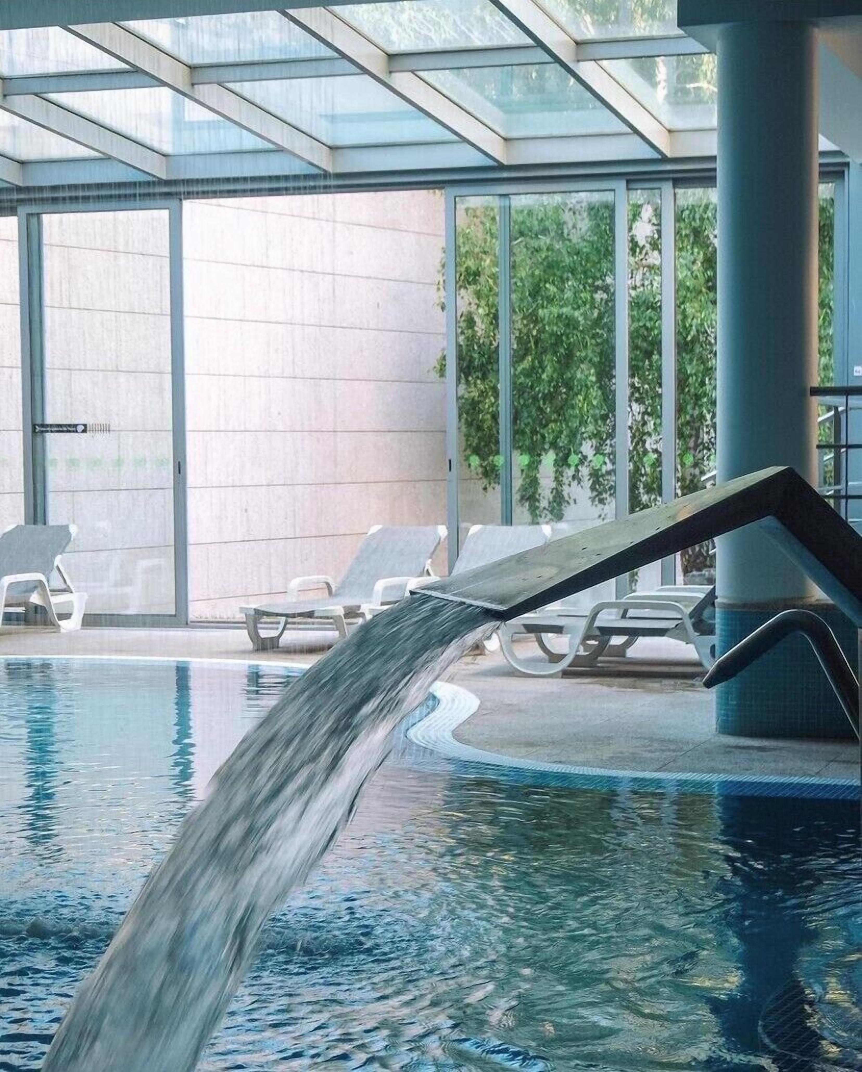 An indoor swimming pool with a waterfall feature, surrounded by lounge chairs and large windows.