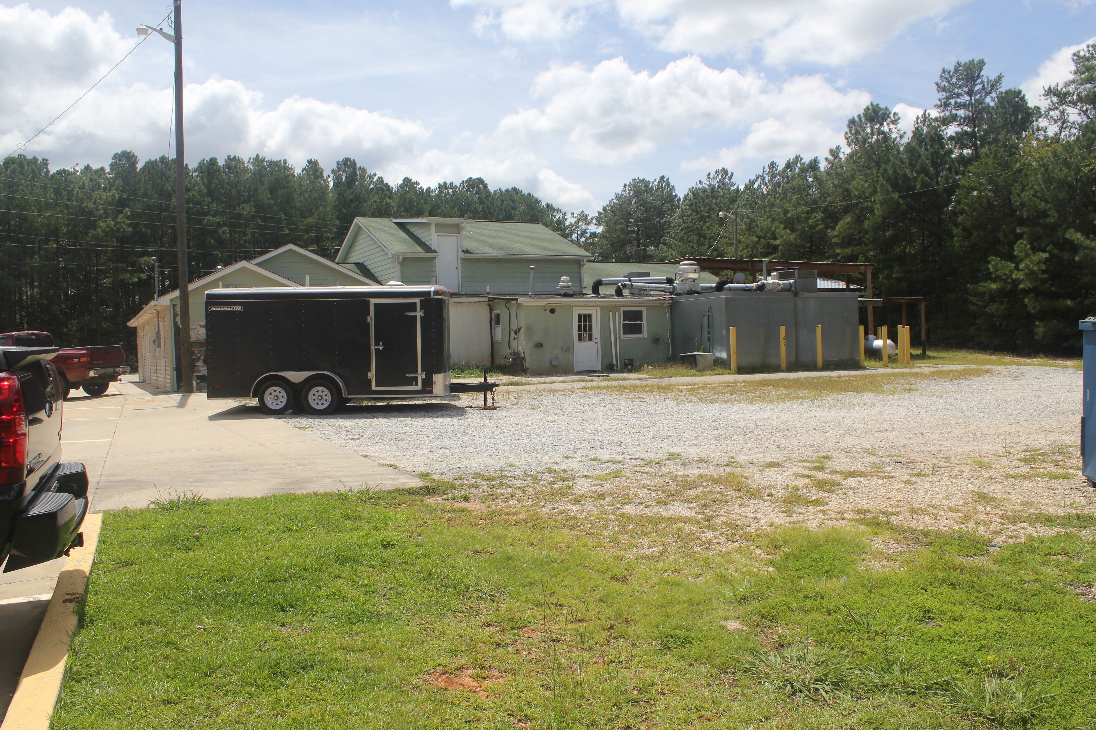 A trailer with a small attached building, a gravel lot, and a wooded area in the background.
