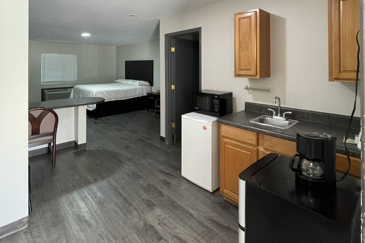 A compact kitchen with a refrigerator, microwave, and coffee maker. There is a bed and a desk in the adjacent room.