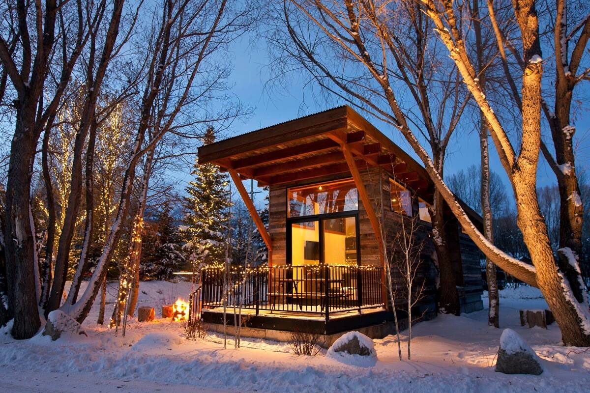 A wooden cabin with a glass front, surrounded by snow-covered trees and a deck with lights.