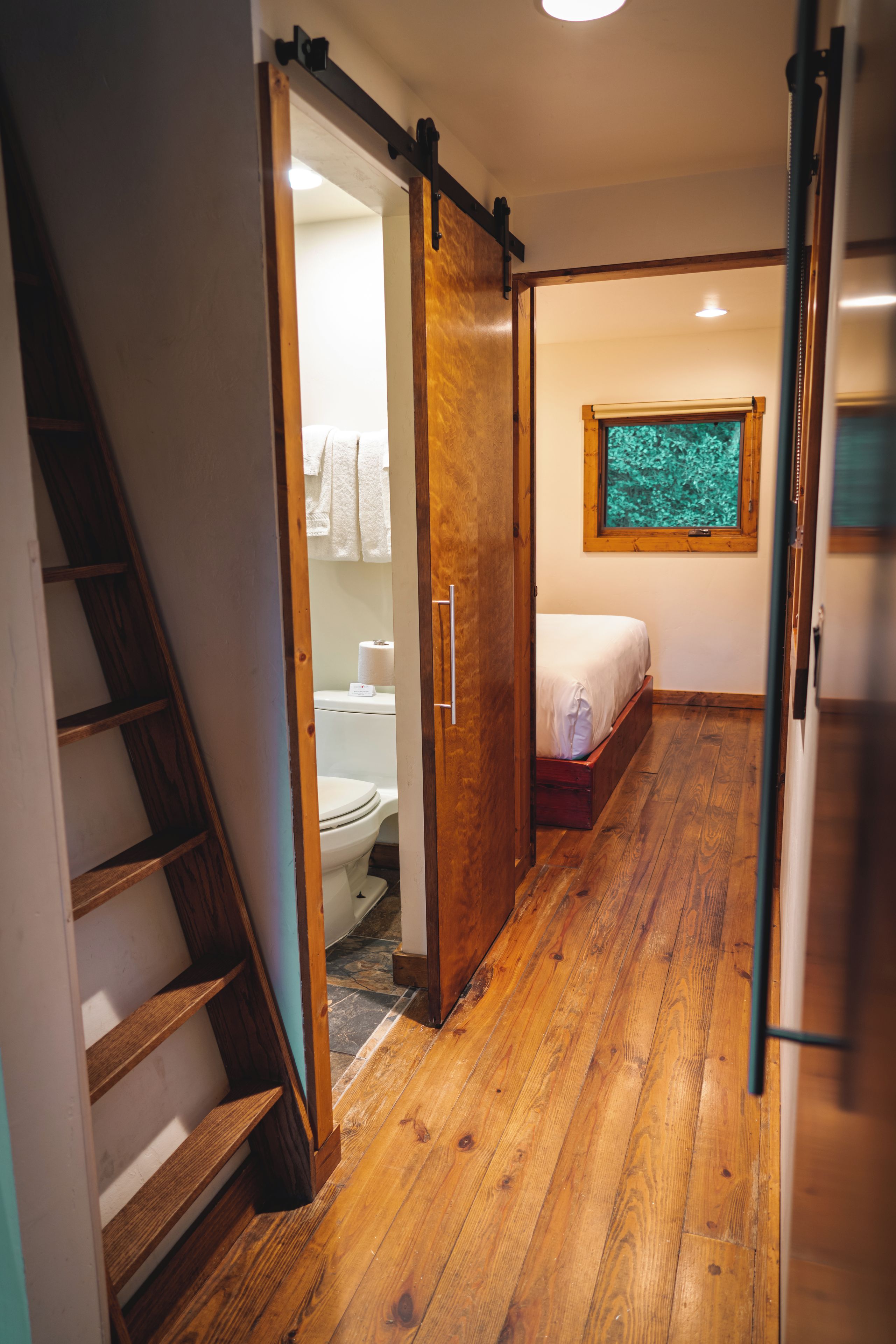 A narrow hallway with a wooden staircase on the left, a toilet with a towel rack in the background, and a bed with a window in the far end.