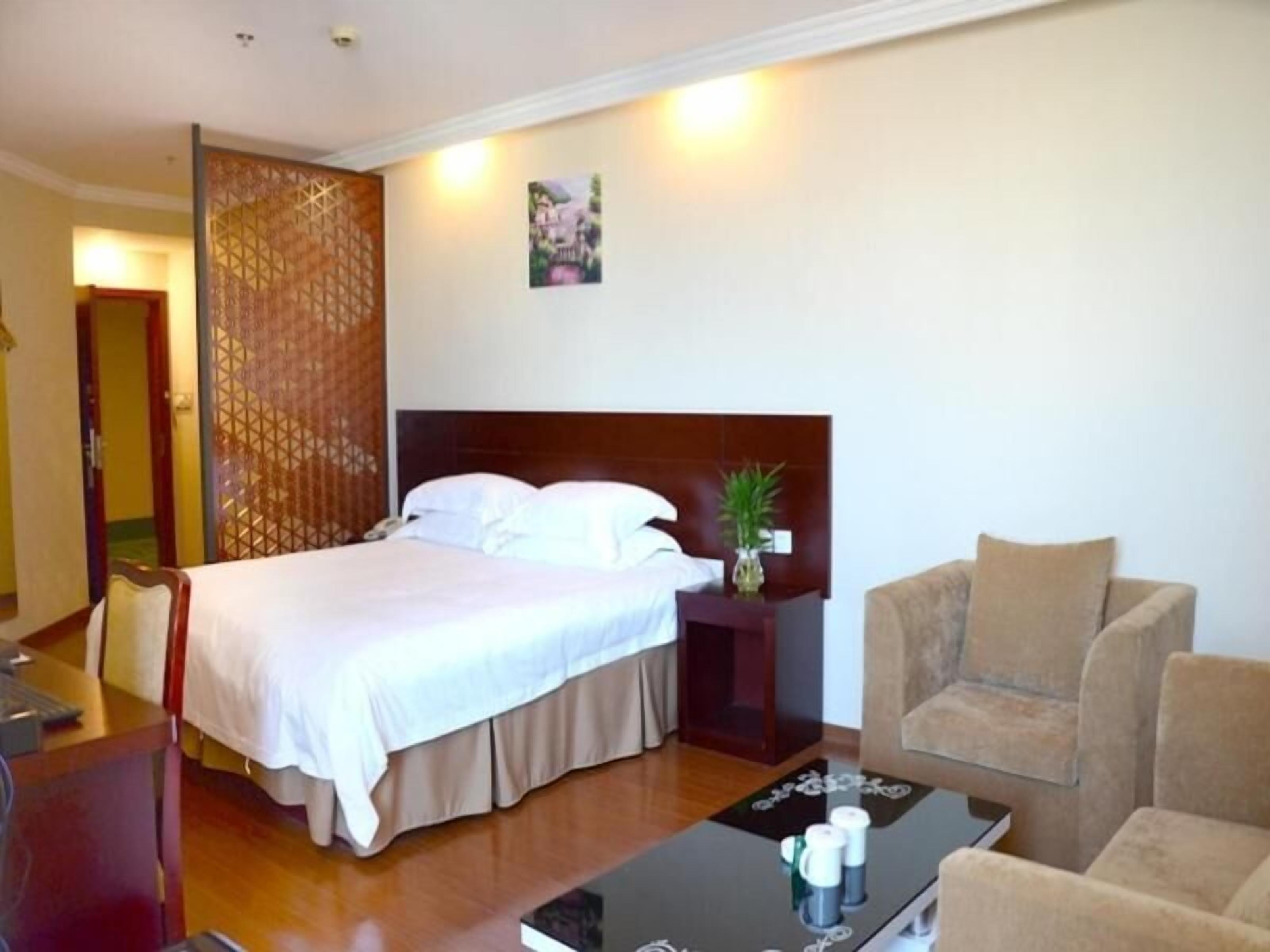 Quarto de hotel com cama, mesa de cabeceira, cadeira, mesa de centro e planta decorativa.