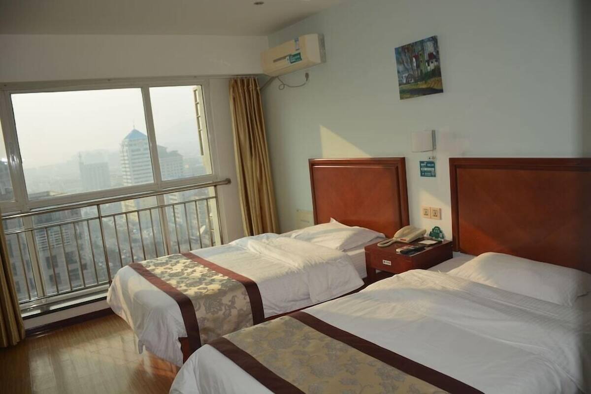 A hotel room with two beds, a window with a view of buildings, a telephone, and a wall-mounted air conditioner.