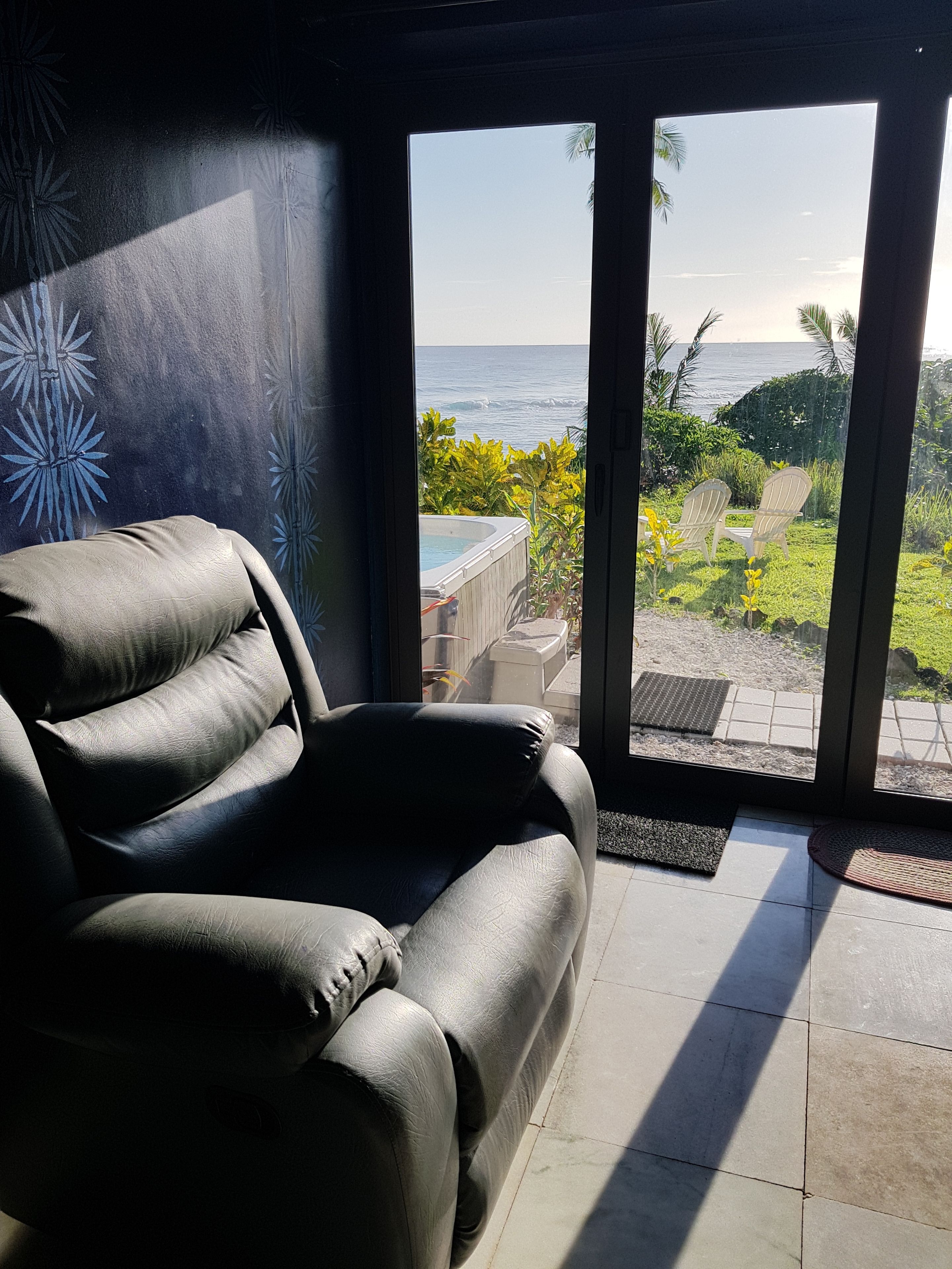 A room with a leather sofa, a glass door leading to a patio with a pool and chairs, and a view of the ocean.