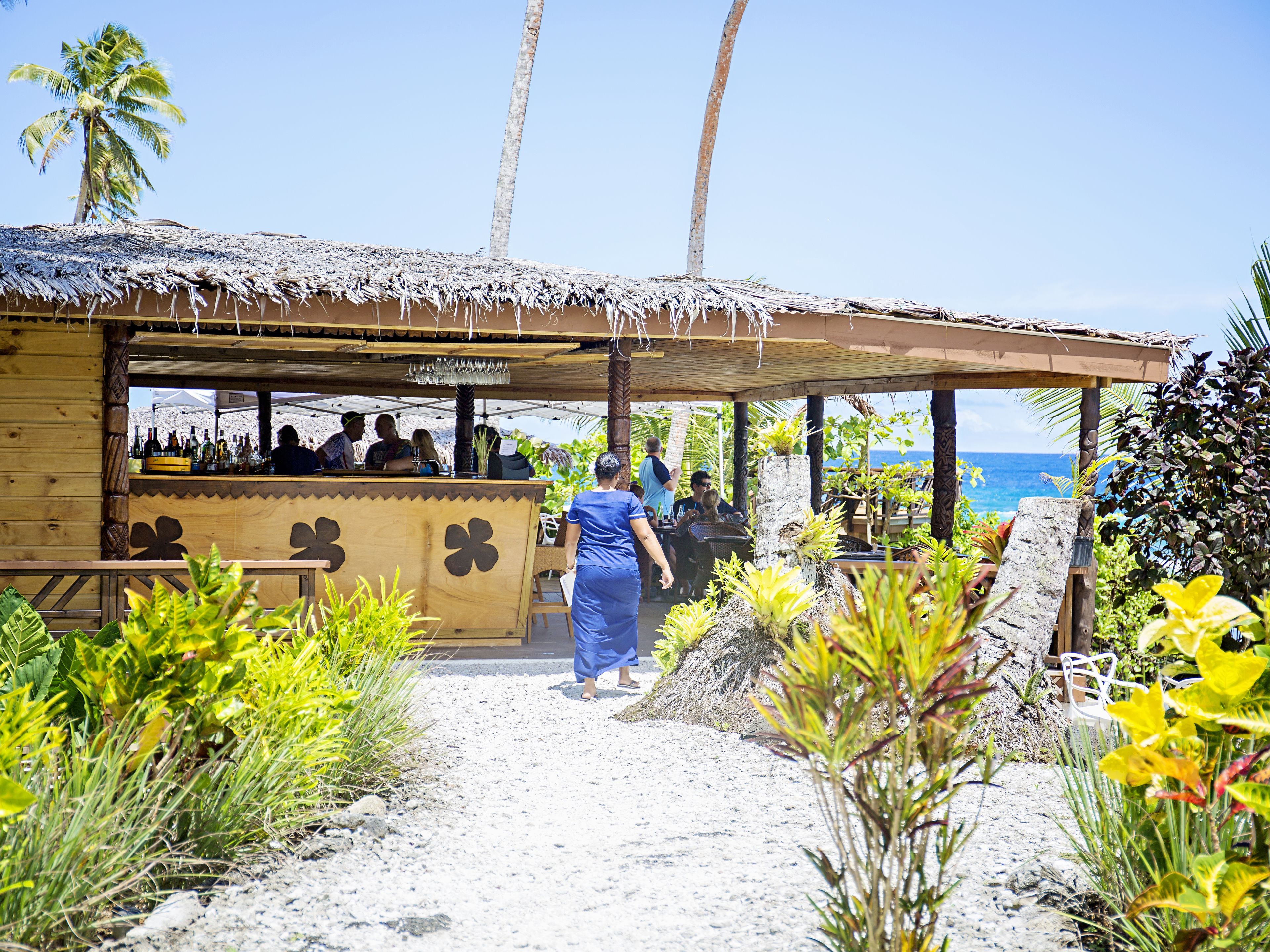 A beachside bar with a thatched roof, surrounded by tropical plants and palm trees, overlooking the ocean.