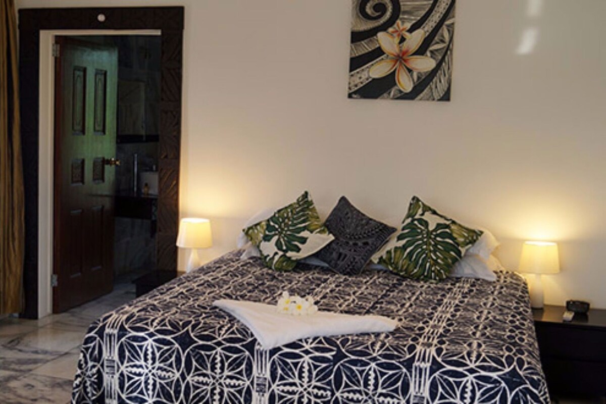 A bedroom with a patterned bedspread, two pillows, a bedside table with a lamp, and a door leading to a bathroom.