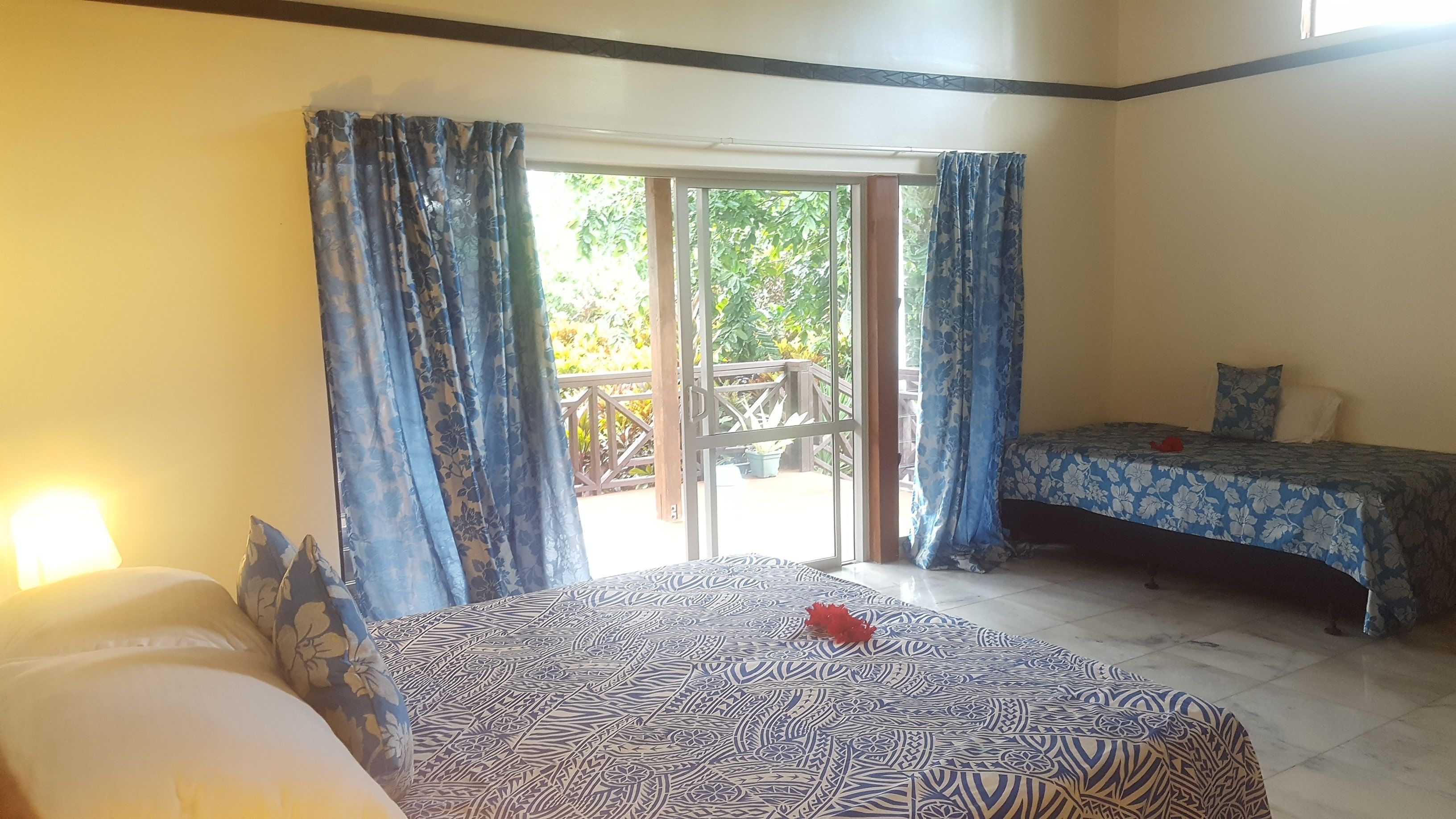A room with two beds, one with a patterned bedspread and the other with a floral bedspread. There is a window with blue curtains, and a door leading to an outdoor area with a wooden deck.