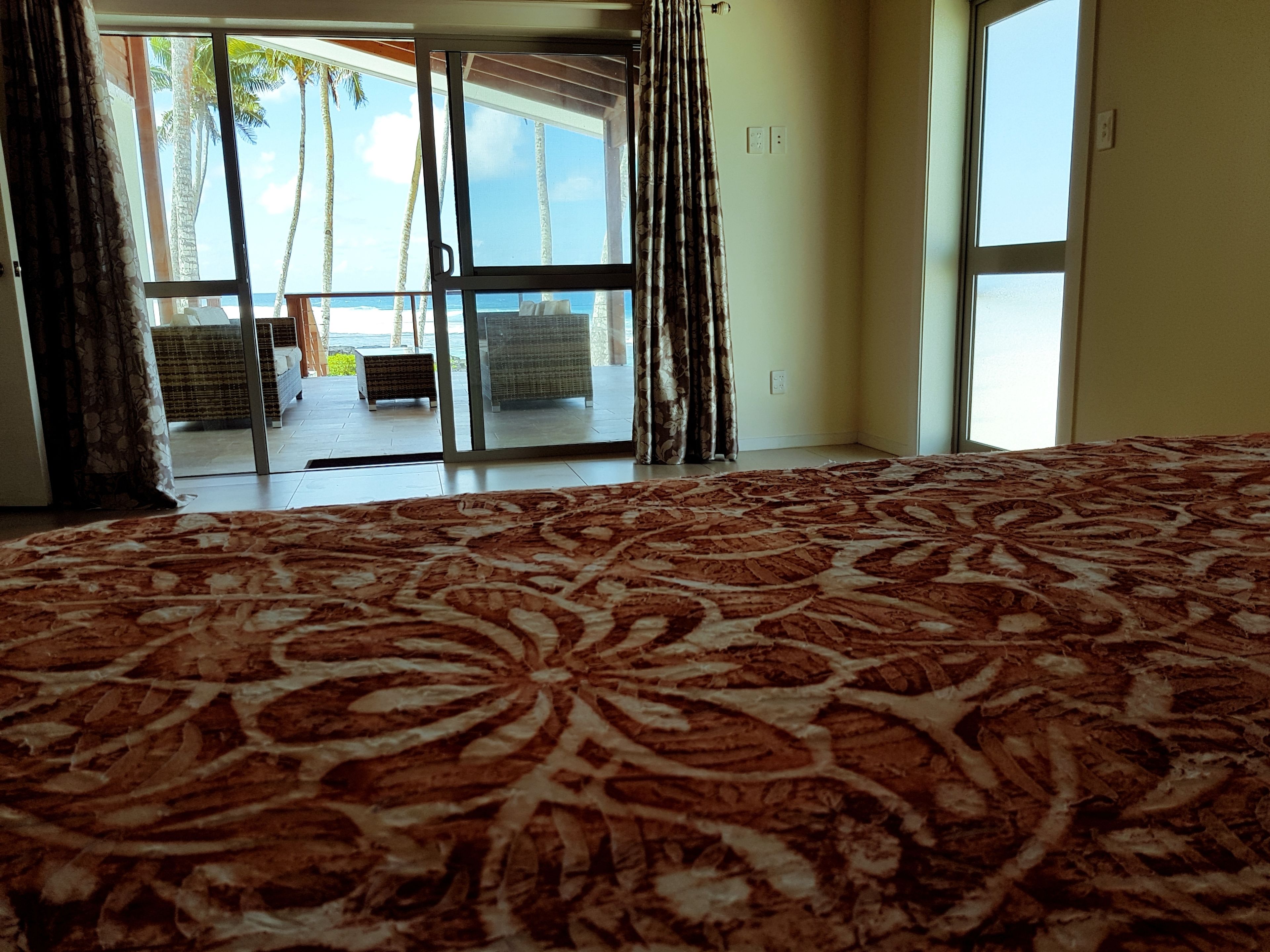 A bedroom with a large bed, a view of the ocean through a sliding glass door, and a patio area with outdoor furniture.