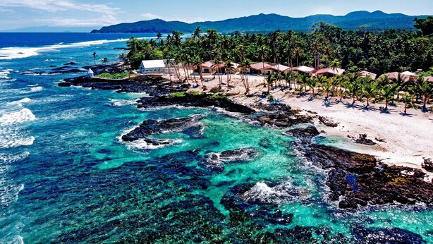 Un resort costero con cabañas de playa, aguas turquesas cristalinas, costa rocosa y montañas al fondo.