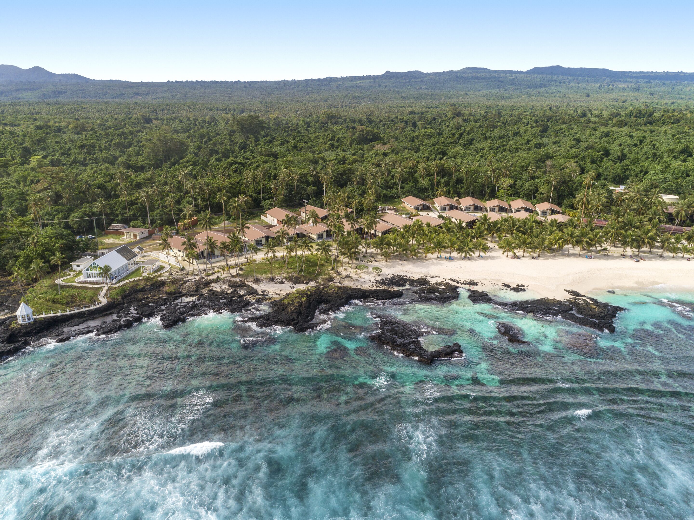 An aerial view of a coastal resort with multiple bungalow-style buildings, a sandy beach, and a rocky shoreline with clear turquoise waters.