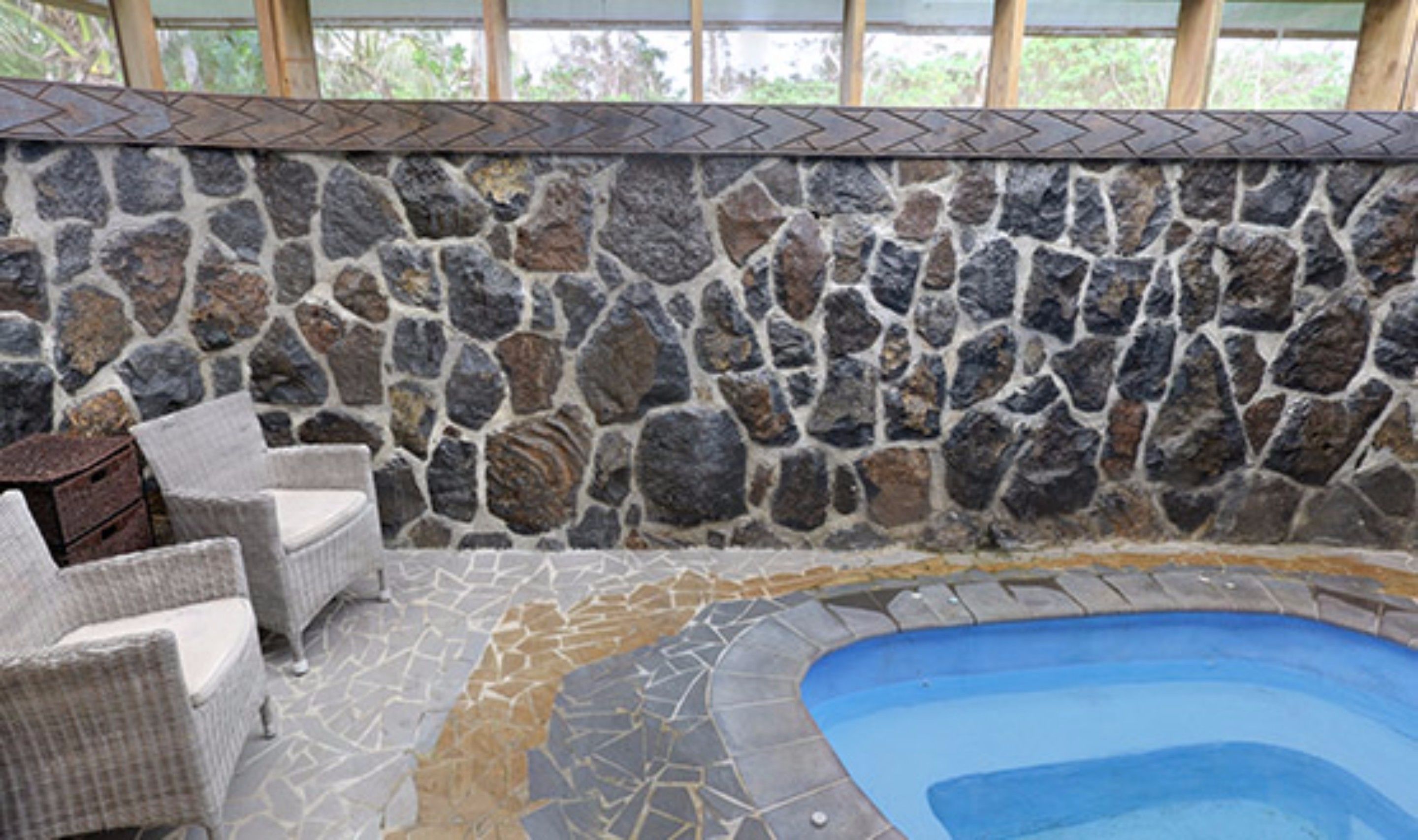 Un área de piscina interior con paredes de piedra y suelo de baldosas, rodeada de sillones de mimbre.