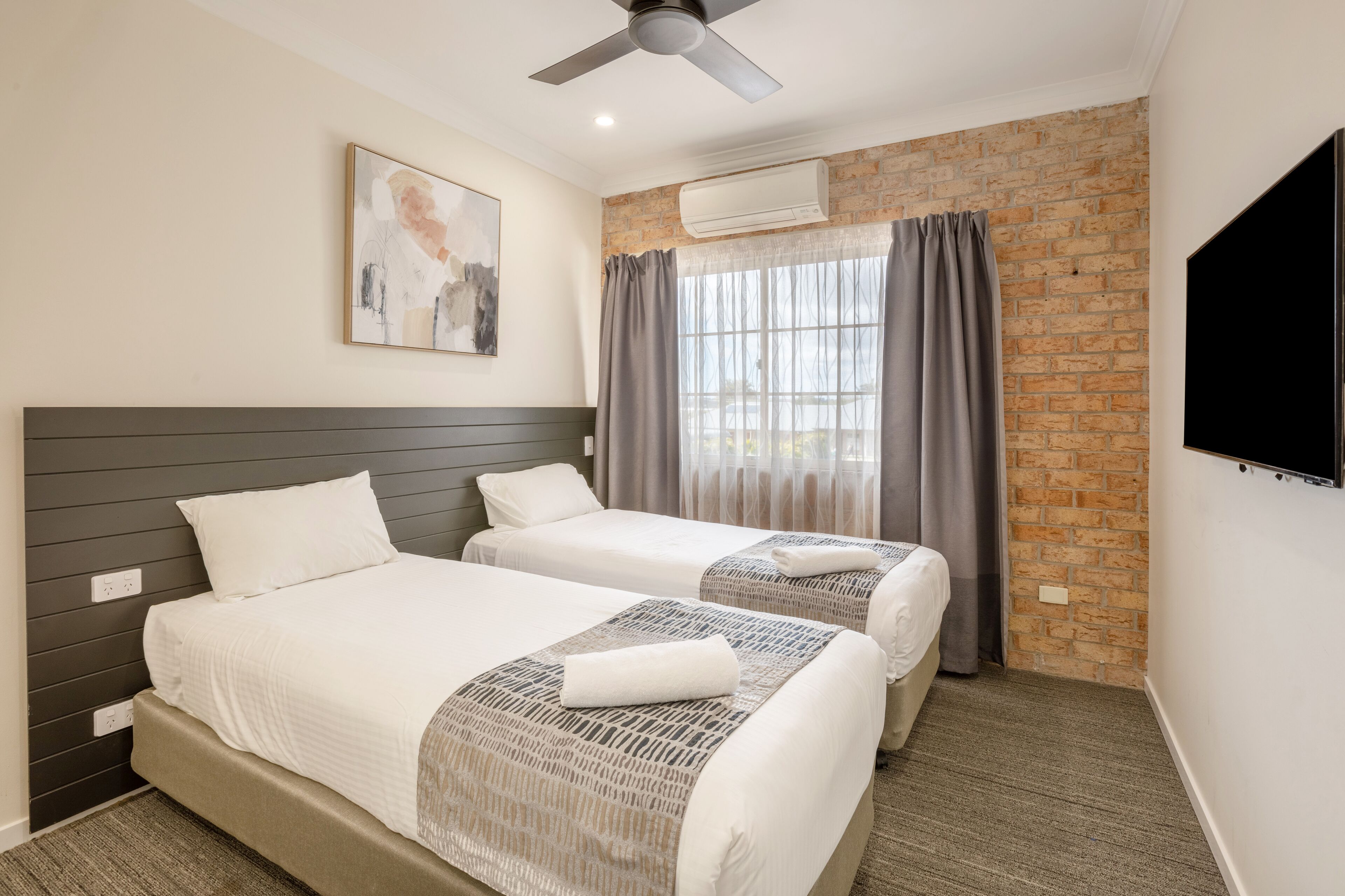 A hotel room with two beds, a ceiling fan, an air conditioning unit, a large window with curtains, and a flat-screen TV.