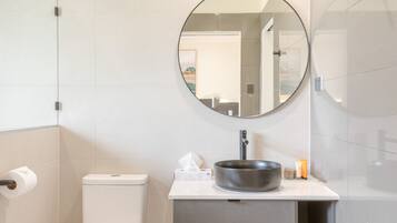 Un baño moderno con inodoro, lavamanos con recipiente grande, espejo redondo y dispensador de toallas.