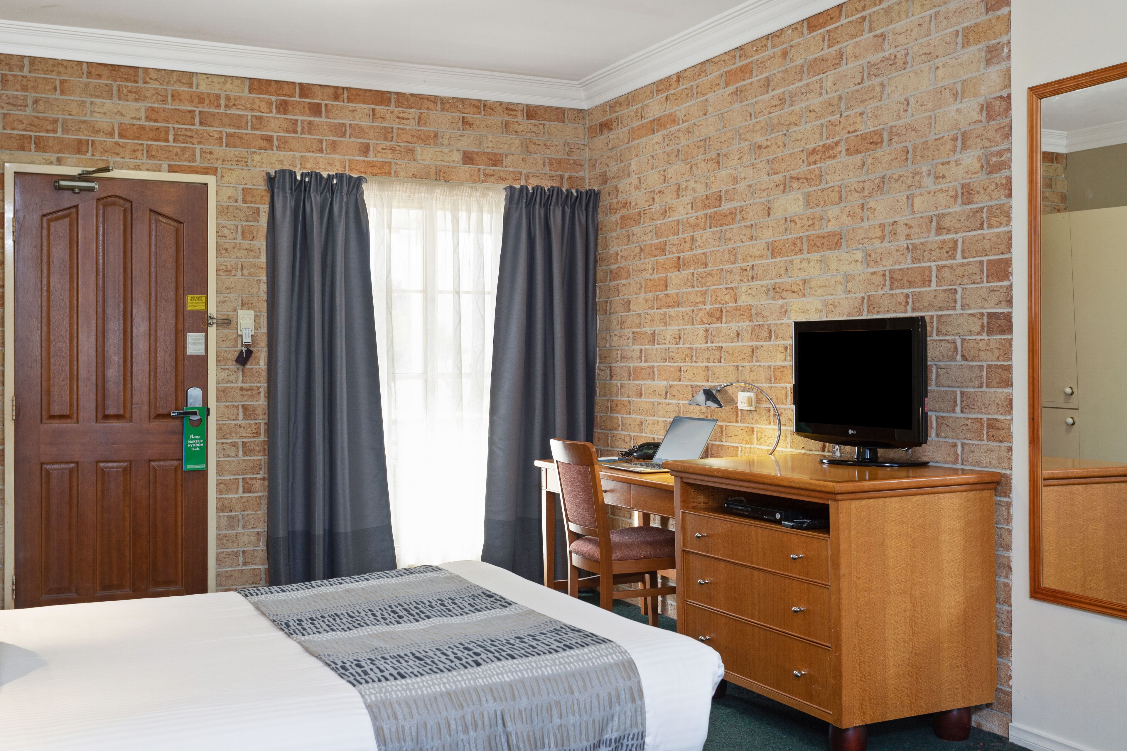 Une chambre d’hôtel avec un mur en briques, un bureau en bois sur lequel se trouve un ordinateur portable, une télévision, un lit recouvert d’une courtepointe à motifs et une porte avec une enseigne verte.