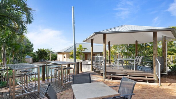 An outdoor patio area with tables and chairs, a covered seating area, and a pool in the background. - Coopers Colonial Motel (Acacia Ridge)