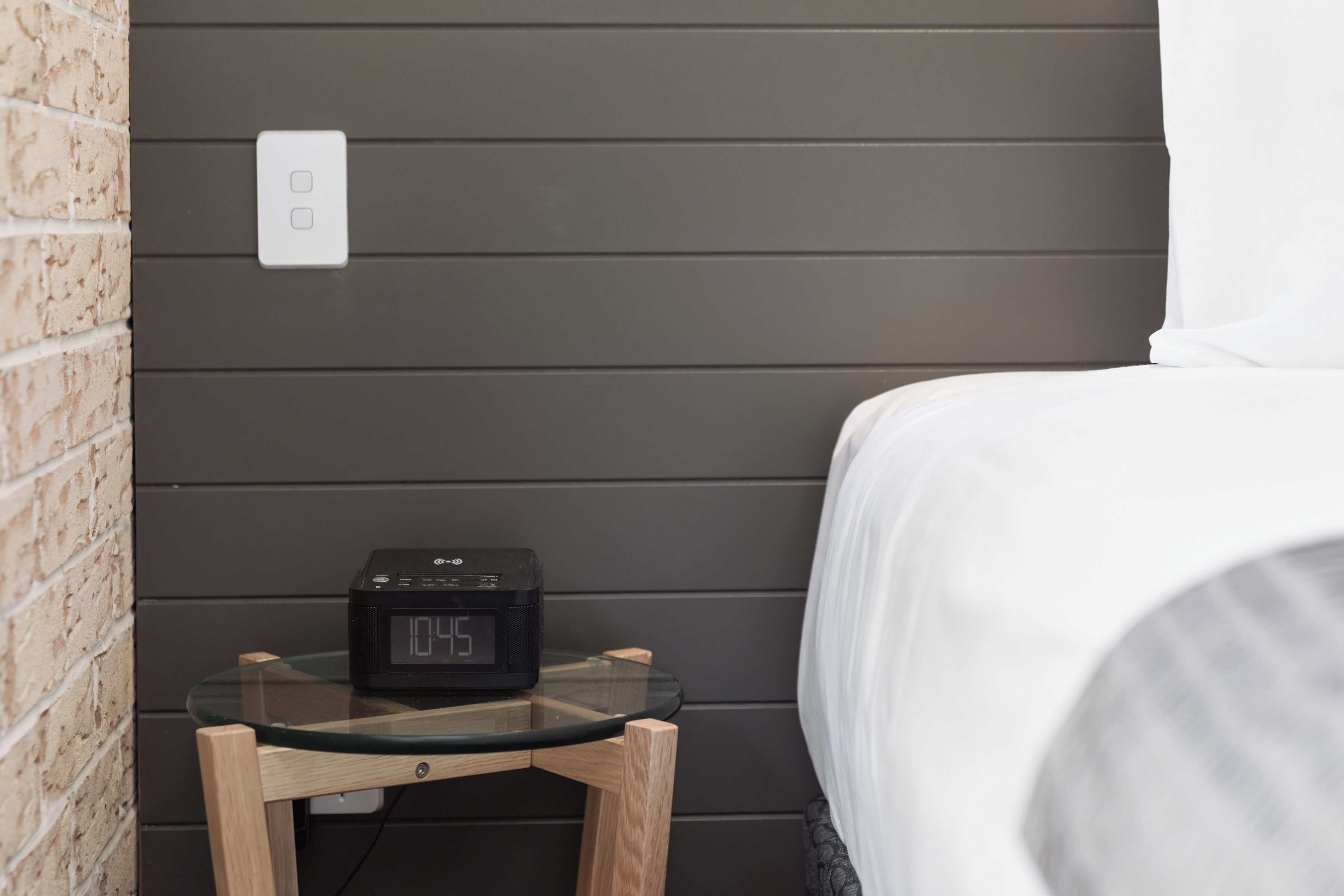A bed with white bedding, a bedside table with a digital clock, and a brick wall.