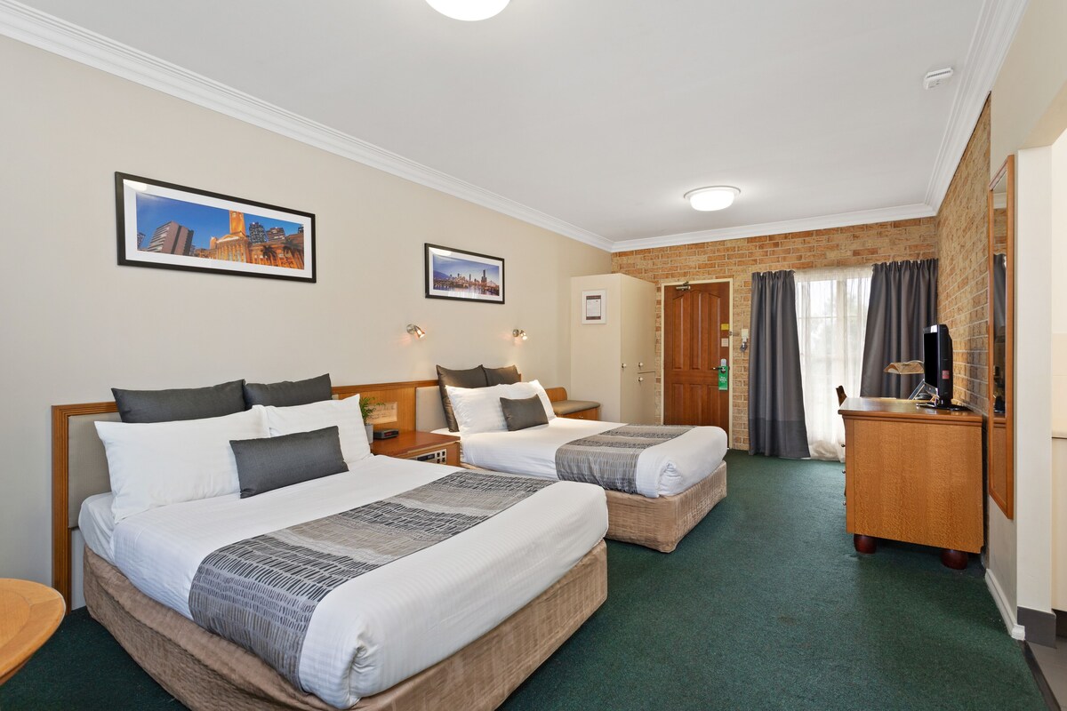 A hotel room with two beds, a desk, a mirror, and a brick wall.
