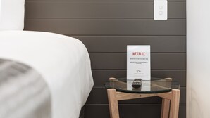 A neatly made bed with a white comforter, a small glass table with a sign for Netflix, and a wall-mounted light switch. - Coopers Colonial Motel (Acacia Ridge)