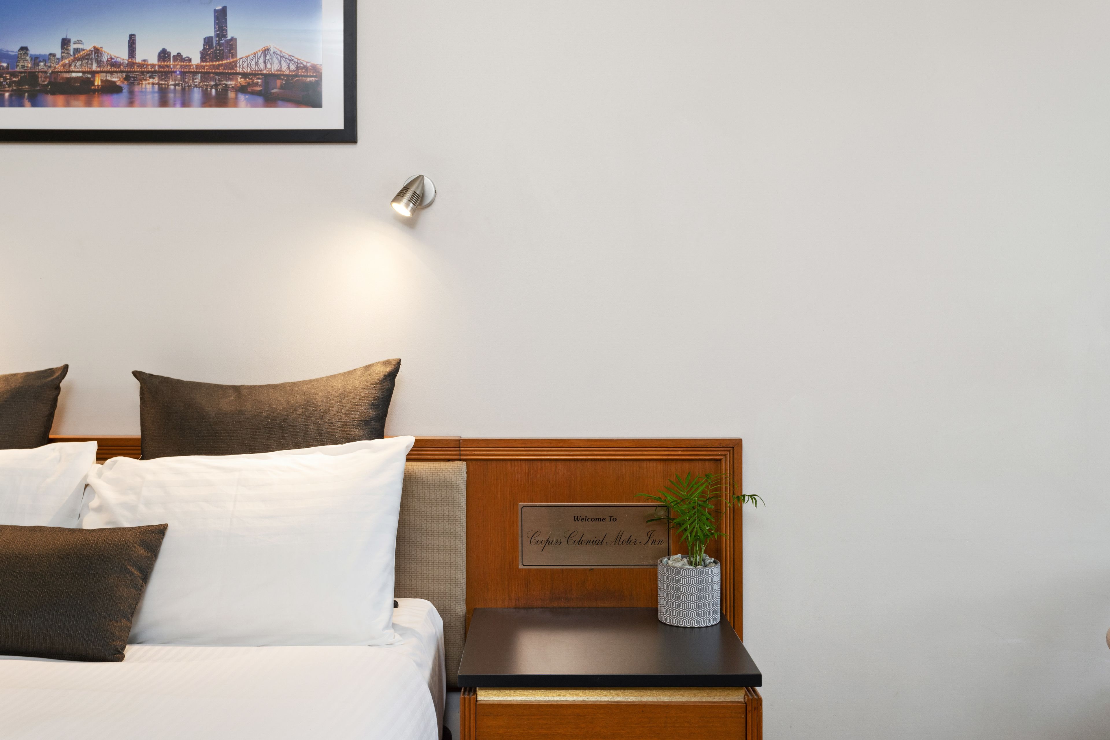 Ein Hotelzimmer mit Bett, Kissen, einem Nachttisch mit einer Willkommenskarte, einem gerahmten Bild an der Wand und einer Zimmerpflanze.