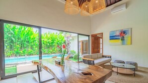 Amplia habitación con una gran mesa de madera, una mesa de centro de vidrio y un área de descanso con sofá y sillones. Un amplio ventanal ofrece vistas a una piscina y exuberante vegetación.