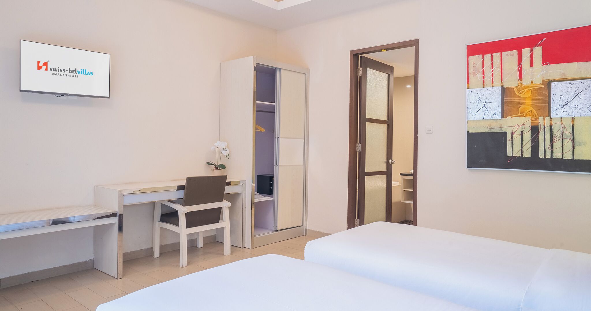 Habitación de hotel con cama, escritorio, silla, armario y televisor que muestra el logotipo de Swiss-Bellvillas.