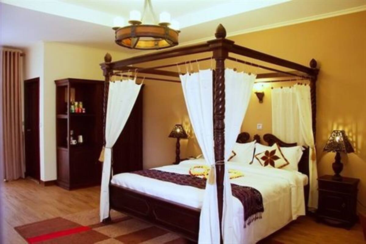 A four-poster bed with white bedding and a canopy, a nightstand with a lamp, and a wooden wardrobe in a hotel room.
