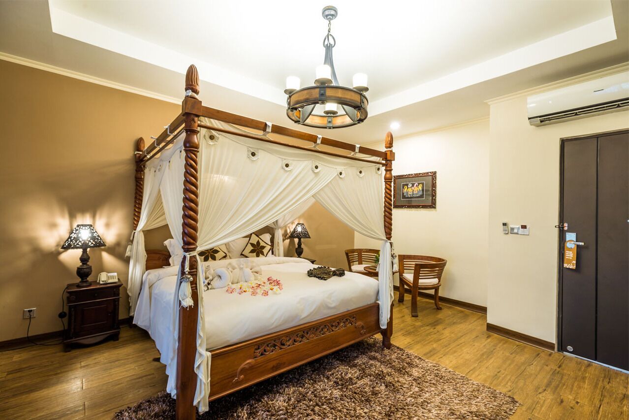 A four-poster bed with a canopy, a chandelier, a wooden floor, a small table with a lamp, and a door with a lock.