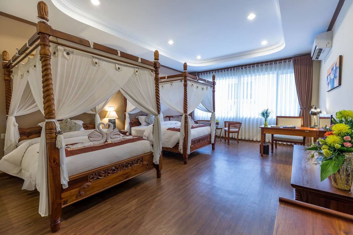 A four-poster bed with white linens and a canopy, a wooden desk with a vase of flowers, and a chair in a hotel room with wooden flooring and a large window with curtains.