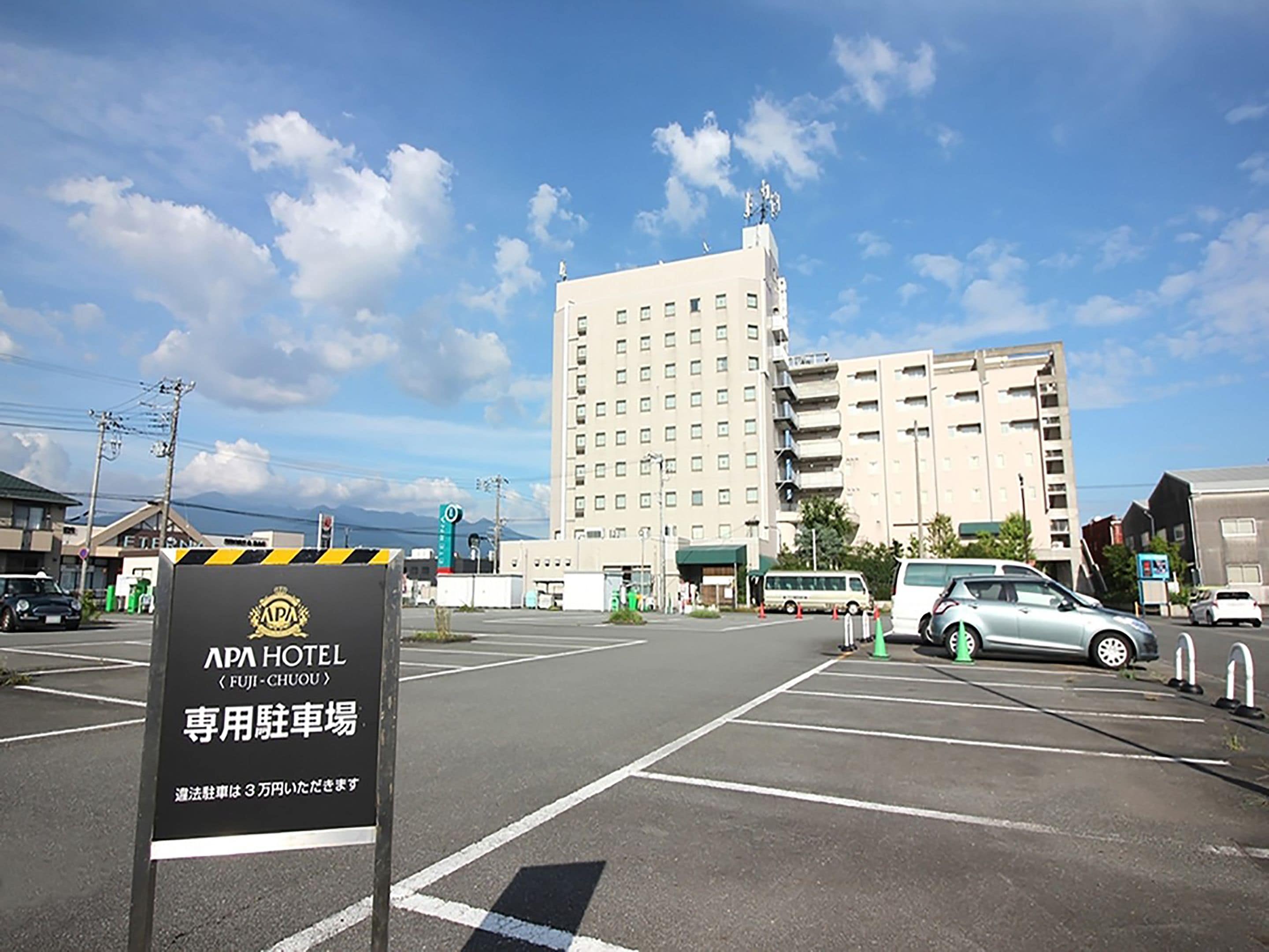 A parking lot with a sign for APA HOTEL (FUJI-CHOUOU) and a large building in the background.