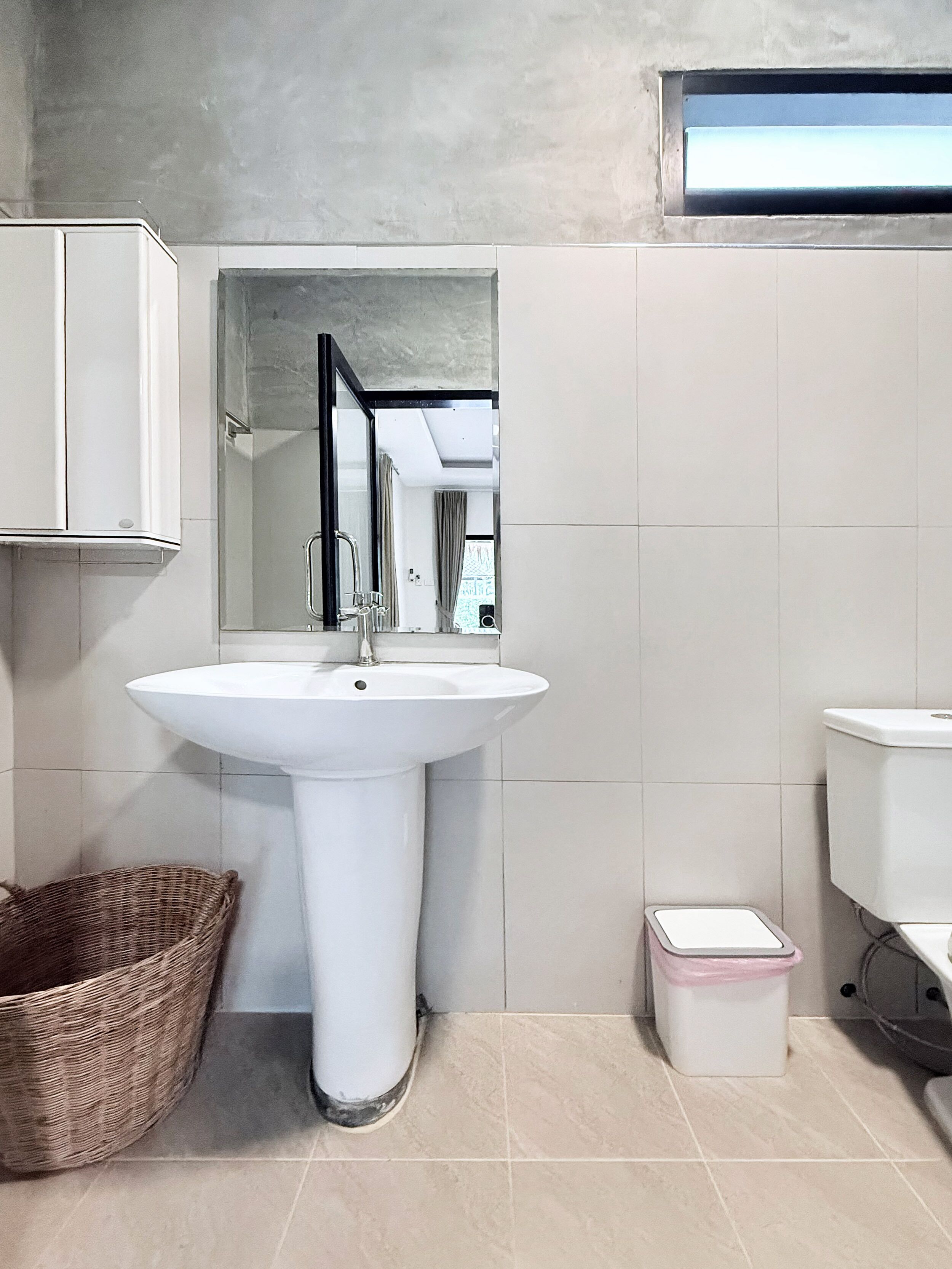 Un baño con un lavamanos blanco, un espejo, una cesta de mimbre y un cubo de basura.