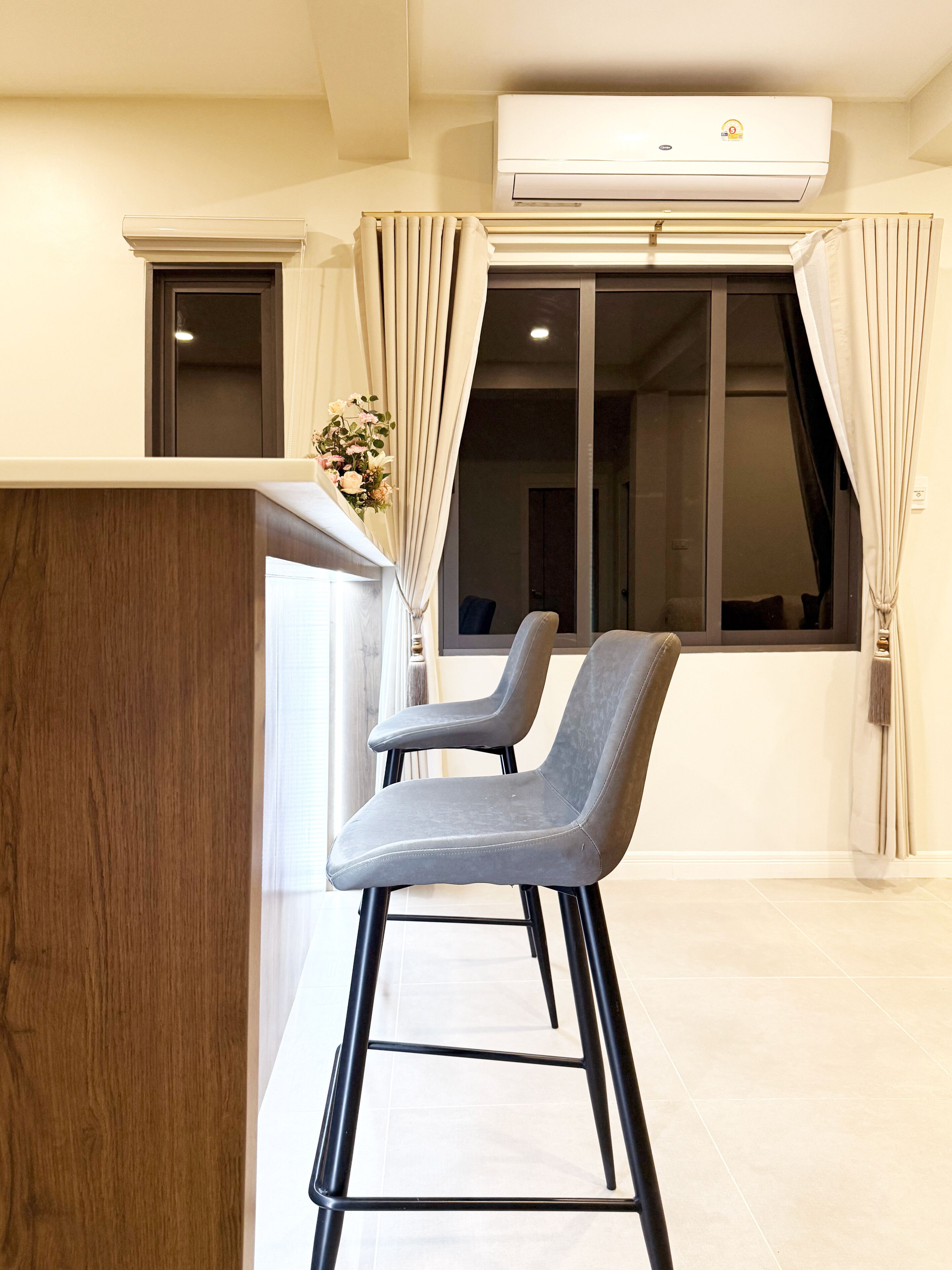 A modern room with a wooden desk, two grey chairs, a large window with curtains, and an air conditioning unit.