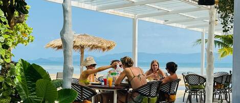 Un espacio comedor al aire libre cubierto con vista al mar, que incluye una mesa con sillas y plantas en macetas.