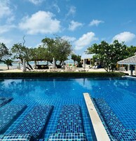 Una piscina con baldosas azul claro, rodeada de sillas de descanso y sombrillas, con vista a la playa y árboles al fondo.